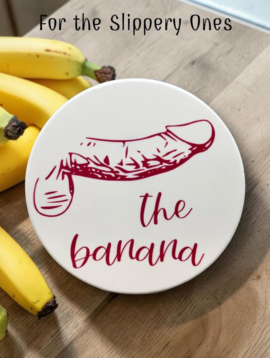 The Banana Dick Type coaster with red penis illustration on a wood counter beside fresh bananas — For the Slippery Ones