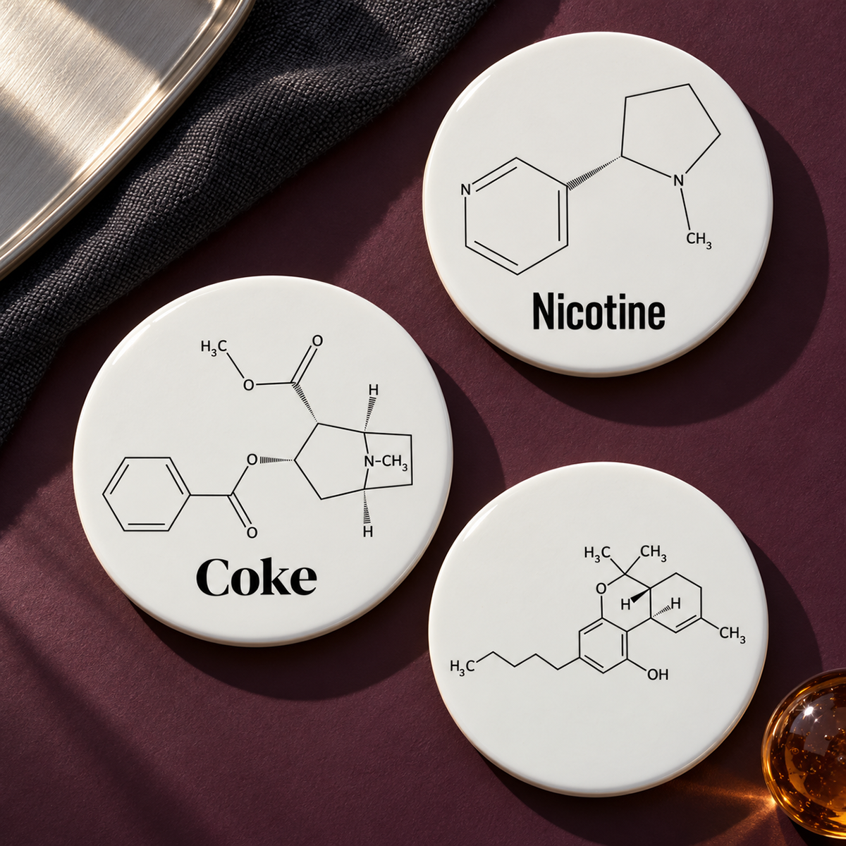 Coke, Nicotine and unlabeled THC molecule ceramic coasters arranged on a plum surface with directional light.
